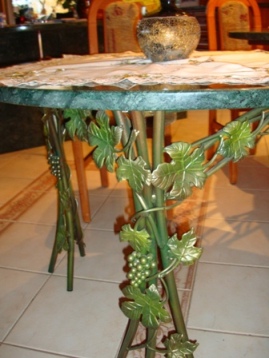 Stół z kutą, metalową podstawą zdobioną motywem winogron i liści, blat z zielonego marmuru, w tle fragment wnętrza.